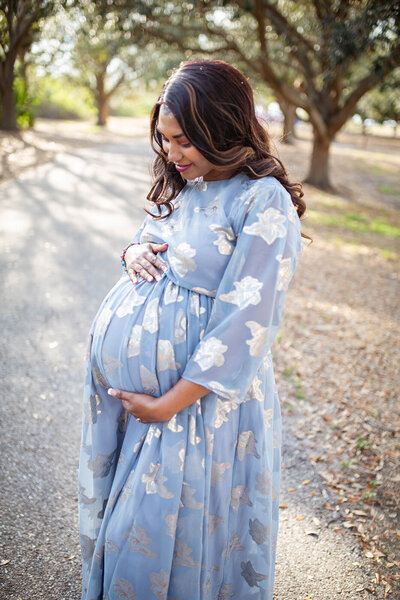 Dark haired woman holding pregnant belly in morning light at park 