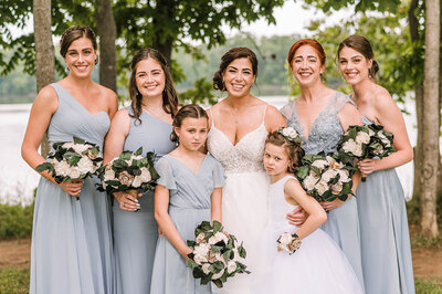 A bride at The Woods at algonkian stands in the middle of her five bridesmaids wearing steel blue and holding bouquets in front of a lake