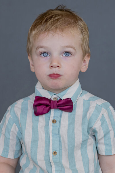 Preschool boy with blue eyes, blonde hair, blue and white striped button down top and a maroon bow. He looks up towards the camera for his school photo.