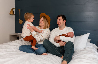 mom and dad sitting on bed and holding newborn baby while playing with toddler