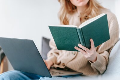 Une jeune femme est assise avec son ordinateur sur ses genoux, elle a dans sa main gauche un carnet ouvert à la couverture verte qu'elle est en train de lire