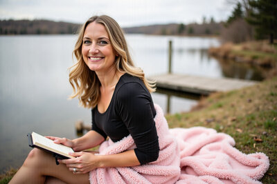 Carissa reading a book by the lake, wrapped in a cozy blanket.