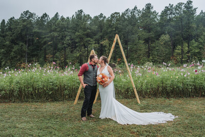 A happy wedding couple shares a romantic kiss in front of a rustic wooden arch adorned with flowers, surrounded by a field of wildflowers and trees at Southern Grace Weddings and Events venue, captured by Quinn Photography LLC.