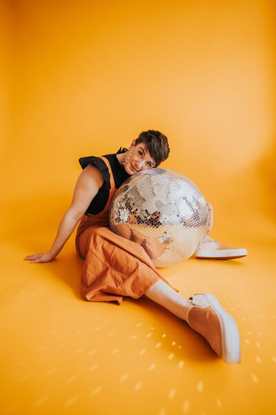 Woman with very short brown hair wears an orange jumpsuit and lays on a disco ball while smiling at the camera in front of a yellow-orange backdrop.