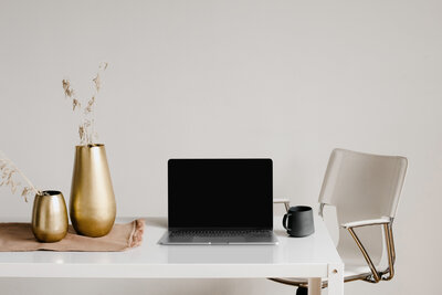  Minimalist desk setup with a laptop, gold vases, and a clear chair in a bright office space.
