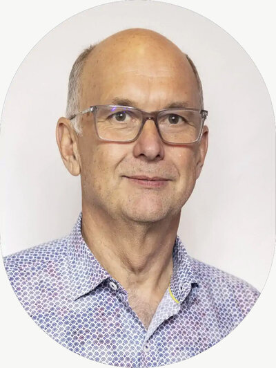 Portrait of an older man wearing glasses and a patterned button-up shirt, smiling slightly against a light background.