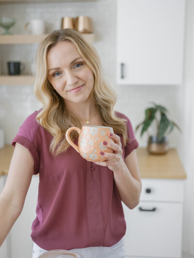 A woman holding a coffee mug in a kitchen smiling and looking at the camera during her brand photoshoot with Krista Marie