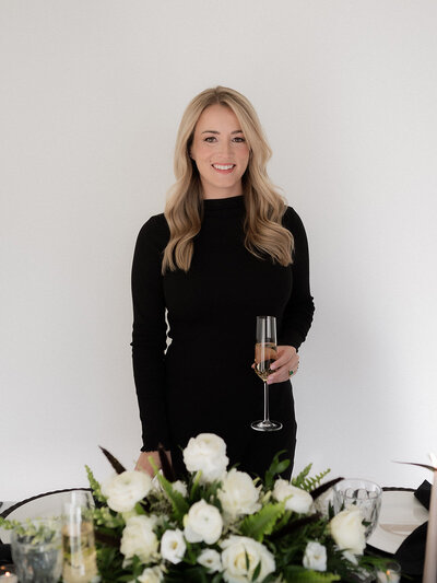 blonde female holding a champagne glass smiling at the camera standing in front of a decorated wedding table