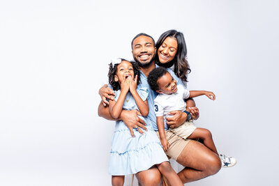 A joyful Black family—mother, father, daughter, and son—laugh together in the studio, dressed in denim and white. A heartwarming moment that captures love and happiness.