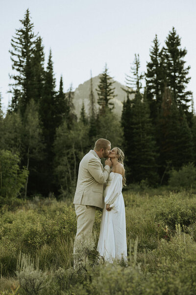 A bride and groom kissing in a green field with a mountain in the distance in Big Cottonwood Canyon, Utah.
