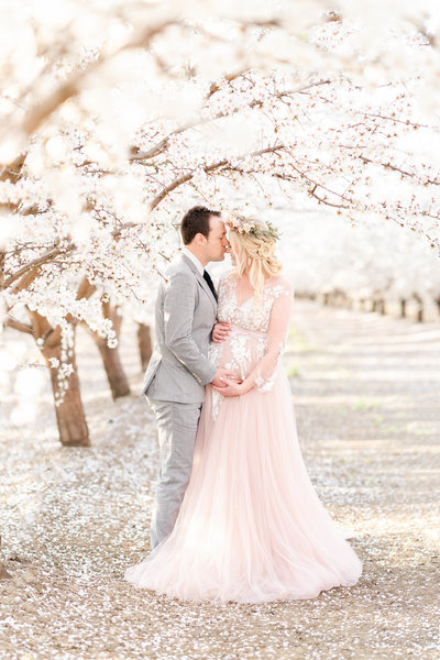 An expecting couple embraces and together hold  mama's belly standing in an almond blossom field during their Bay area maternity photography session by Light Livin Photography