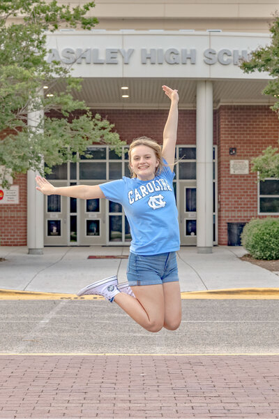 A graduating senior in a University of North Carolina blue shirt and shorts joyfully jumping in front of Ashley High School in Wilmington North Carolina. This energetic image captures the excitement of senior year, ideal for capturing vibrant and fun senior photography.