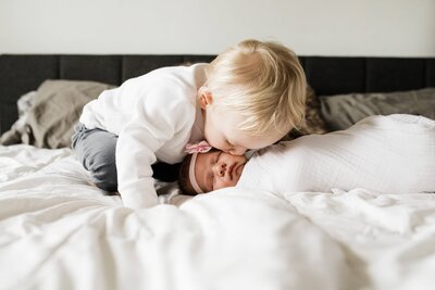 A toddler gently kissing a newborn who is lying on a bed during an in-home newborn photography session.