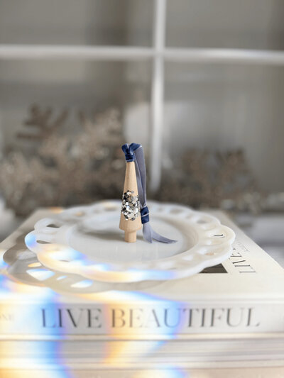 A decorative party favor resembling a holiday ornament, adorned with ribbon and sequins, sits on a white plate atop a large book titled "LIVE BEAUTIFUL." Design inspiration by Sweet Pea Ink Creative.