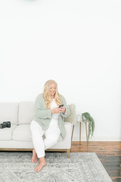 Deborah Jossick, owner of Oak & Lore Studio, sits on a cozy couch in her studio, looking at her phone with a thoughtful expression, surrounded by the creative space where she brings her clients' stories to life.