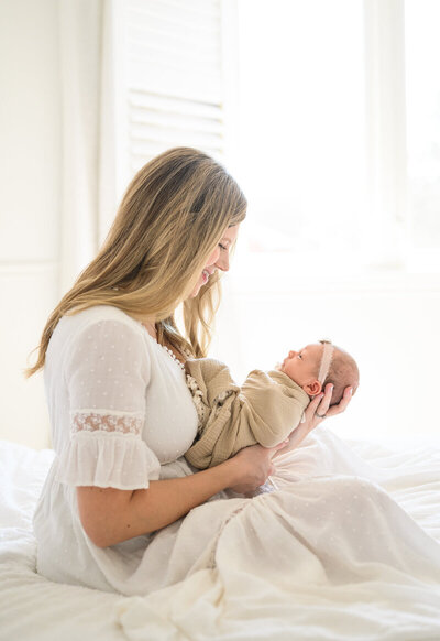 A woman wearing a white dress sits on a bed, gently holding a swaddled newborn baby in her arms, both smiling, embodying the elegance you'd expect from showit web design for luxury photographers.