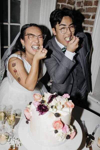 A bride and groom laughing and feeding each other cake at their reception. The bride's arm tattoos are visible, and the table is adorned with a floral cake and champagne glasses.