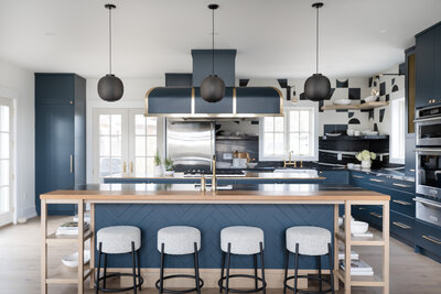 A wood and stone kitchen island lined with plush beige stools  and featuring open shelves and deep blue decorative paneling