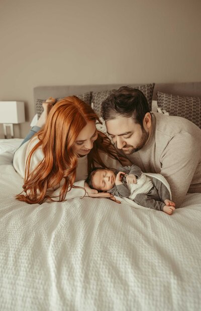 Newborn photo session with parents at home. The loving parents of the newborn baby are lying on a bed with white and pastel brown blankets. Their faces light up with big smiles as they look at their precious baby, creating an intimate and heartwarming scene
