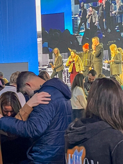 Nita Tin standing on stage at Passion City Church in Atlanta, holding a microphone and praying alongside Aaron Coe. The Passion City Band is in the background, with congregants in the foreground praying with hands on one another’s shoulders. She wears a red dress and jacket.