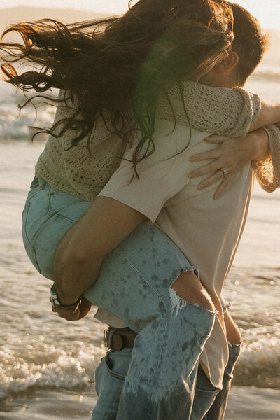 a woman bear hugging a man on the beach during sunset