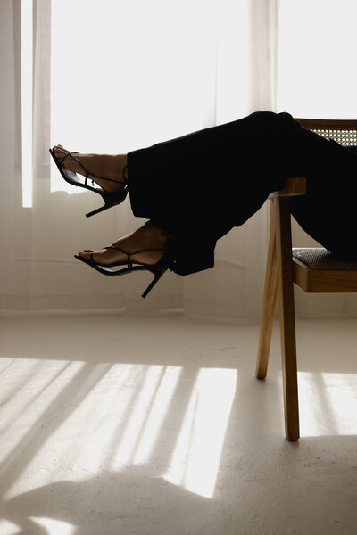 A woman's legs hanging from a chair in a black suit and black high heeled sandals