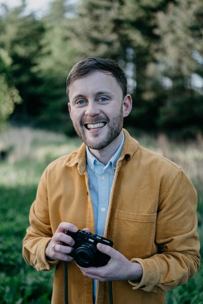 Aberdeen wedding photographer Scott Arlow stands in woodland wearing a yellow shirt.