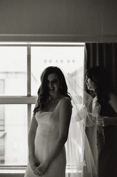 Bride in front of window with maid of honor putting her veil on