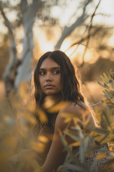 A woman with long, wavy hair looks to the side, surrounded by foliage in a sunlit outdoor setting, embodying her brand identity amidst nature's serene beauty.