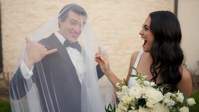 Raegan holds up a hang loose sign in his black tux and black bowtie with his bride's veil covering him. Faith playfully looks shocked and points at him as she holds her ivory flora bouquet at their luxury wedding venue in Canyon Lake Texasl