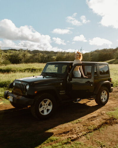 young woman smiles at the camera while stepping into her Jeep  in a field in Hawaii