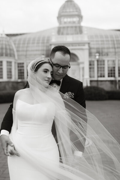 Bride and groom embracing outside the Franklin Park Conservatory in Columbus, Ohio, from Kylie and Thomas’s wedding, photographed by Rainstorm Photo & Video in soft, timeless black and white.