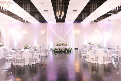 A romantic reception space at SoHo63, a Chandler wedding and event venue, featuring round tables with white linens, silver chiavari chairs, and a sweetheart table framed by flowing white drapery and floral arrangements. Soft purple uplighting and modern chandeliers complete the elegant look.
