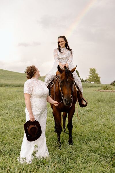 Two brides, one riding a horse, the other holding the horse's bridle. There is a rainbow behind them.