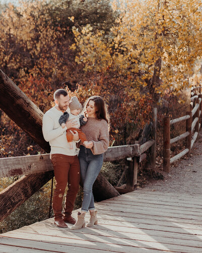 A small family of three smiles together while standing on a bridge surrounded by beautiful autumn colored trees in a Colorado state park.