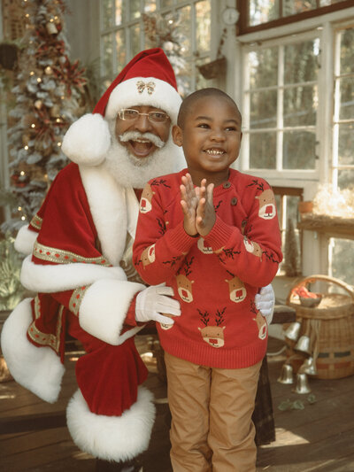 A super excited little boy clapping and standing with The Black Santa of Atlanta.