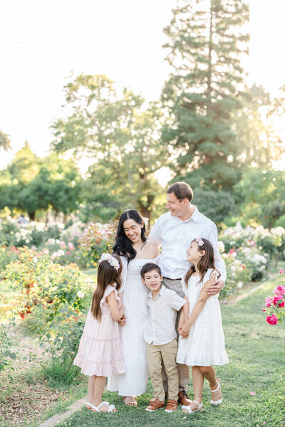 A family of four stand in a field of purple lupines dressed in shades of grey, blue, and white while smiling and holding one another photographed by bay area photographer, Light Livin Photography.