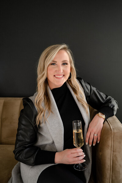 Blonde female smiling and sitting on a beige velvet couch with a champagne flute in her hand with a black wall behind her.