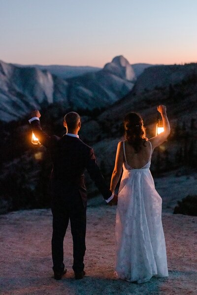 A bride and groom hold lanterns at dusk, while looking at the back side of Half Dome in Yosemite National Park. 