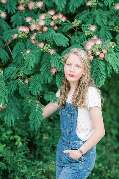 Senior girl in overalls smiling in front of green leaves
