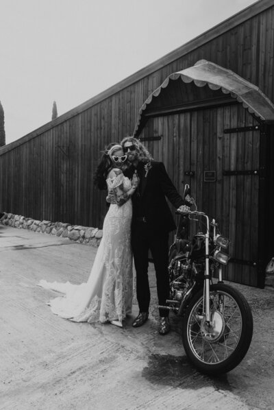 Black and white shot of a cool couple poses confidently next to a motorbike, epitomising their alternative and stylish approach to love and life at Willow Marsh Farm