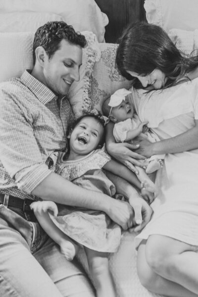 black and white photo of a happy cuddling family laying down in bed with their big sister and newborn. Newborn and toddler girl are between mom and dad and everyone is smiling.