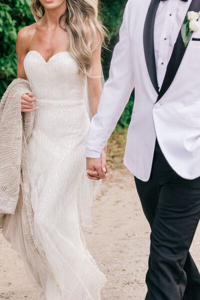 A romantic close-up of a bride and groom holding hands as they walk along a scenic path at their Palmetto Bluff wedding in South Carolina. The bride stuns in a strapless beaded gown, while the groom wears a classic white tuxedo, creating an elegant and timeless wedding moment.