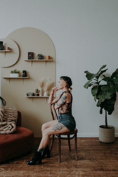Profile view of the photographer sitting on a wooden chair, wearing a lace top and shorts, surrounded by cozy textures and neutral tones.