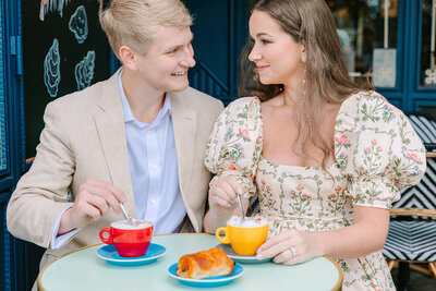 Couple holding coffee cups and gazing at each other inside a colorful Parisian café near Luxembourg Gardens in the Latin Quarter during a romantic photoshoot.