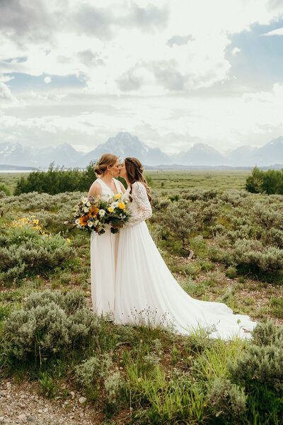 two brides kissing while standing outside in their wedding dresses with mountains in the distance
