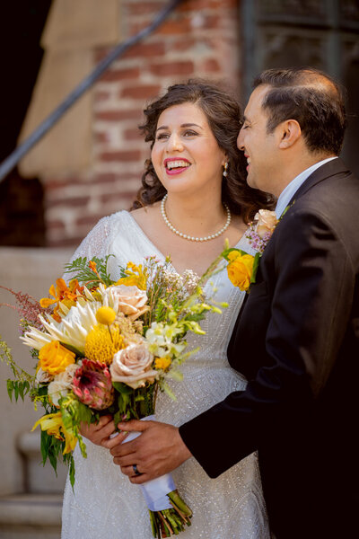 Smiling couple holding a colorful bouquet in front of a brick backdrop