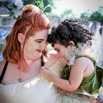 Red-haired woman and curly-haired child sharing a sweet moment in Ren Faire fantasy costumes with braided and tousled hairstyles.