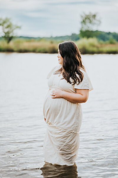 Expecting mother holds belly while standing in water during maternity session in Raleigh NC