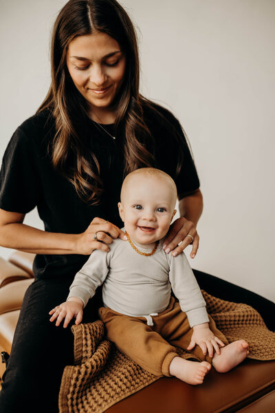 A young brunette, female chiropractor palpates a six month old baby boy's shoulder as he makes direct eye contact at the audience, while sitting up and smiling a huge toothless grin.
